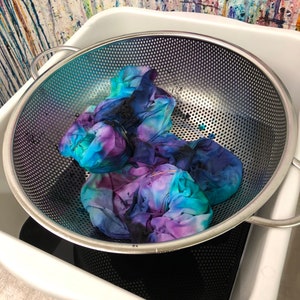 May include: A white plastic tub with a stainless steel colander inside. The colander contains a tie-dye fabric in shades of blue, purple, and black.