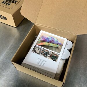 May include: An open cardboard box containing an ice dye kit. The kit includes a white cotton pillowcase, rubber gloves, soda ash, fibre reactive dyes, and instructions. The box is labelled "Wildflower Dyes, Live in Color".