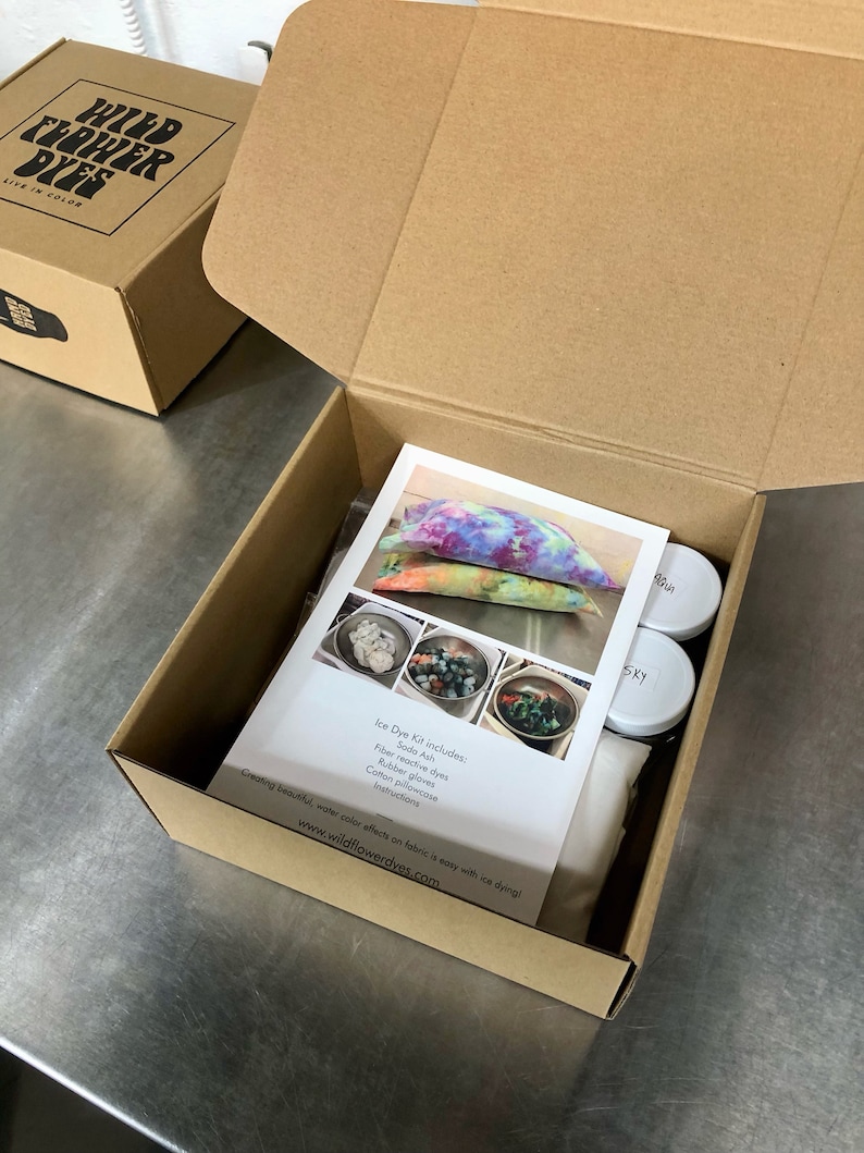 May include: An open cardboard box containing an ice dye kit. The kit includes a white cotton pillowcase, rubber gloves, soda ash, fibre reactive dyes, and instructions. The box is labelled "Wildflower Dyes, Live in Color".