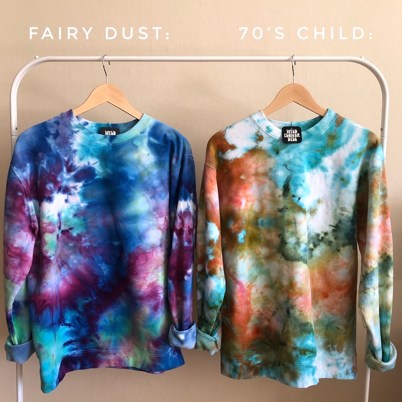 May include: Two tie-dye sweatshirts hanging on a rack. The sweatshirt on the left is blue, purple, and green. The sweatshirt on the right is orange, green, and blue. The text "FAIRY DUST" is above the left sweatshirt and the text "70'S CHILD" is above the right sweatshirt.