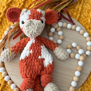 May include: A crocheted plush toy cow with orange and white fur. The cow is sitting on a wooden surface with white and brown beads around it.