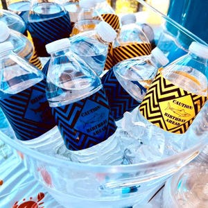 May include: A clear plastic tub filled with ice and water bottles. The bottles are decorated with blue and yellow labels that say "Caution: Birthday Shark Ahead".