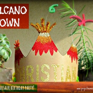 Puede incluir: Una corona de cartón marrón decorada con purpurina roja y dorada, con forma de volcán con una parte superior de purpurina roja y dorada. La corona dice "VOLCANO CROWN" en letras rojas y "CHRISTIAN" en letras de purpurina dorada. La corona es para una fiesta de cumpleaños de dinosaurios.
