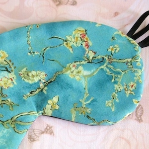 May include: A blue sleep mask with a floral pattern of white blossoms and green branches. The mask has a black elastic strap.