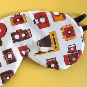 Op de afbeelding: Een wit slaapmasker met een zwart elastiek. Het masker is versierd met een herhalend patroon van vintage camera's in verschillende kleuren, waaronder rood, oranje, bruin en geel.