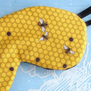 Könnte beinhalten: Eine gelbe Schlafmaske mit einem Wabenmuster und drei kleinen Bienen-Illustrationen.