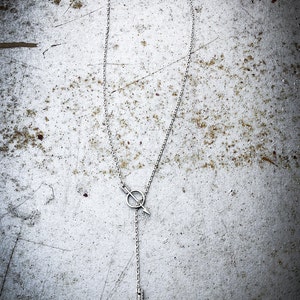 May include: A silver chain necklace with a pendant featuring a circle with an arrow pointing through it. The arrow is attached to a longer chain that ends in a pointed arrow.