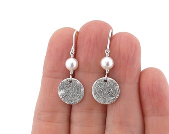 May include: A pair of silver drop earrings featuring a round, textured metal disc with an engraved floral pattern. Each earring is accented with a single white pearl bead suspended from a silver hook wire, creating a balanced design.