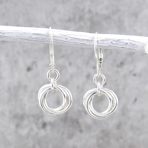 May include: A pair of silver-colored earrings. Each earring features a cluster of interlocking rings suspended from a lever-back closure. The earrings are displayed against a light gray background, creating a minimalist aesthetic.