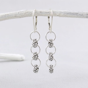 May include: Silver dangle earrings with a chain of four flower-shaped beads. The earrings have a hook closure.