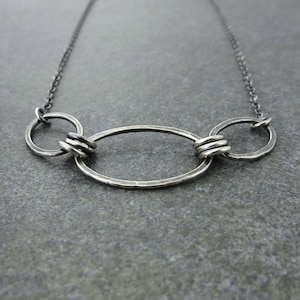 May include: A silver necklace with three interconnected circles. The circles are of different sizes and have a hammered texture. The necklace is on a gray background.