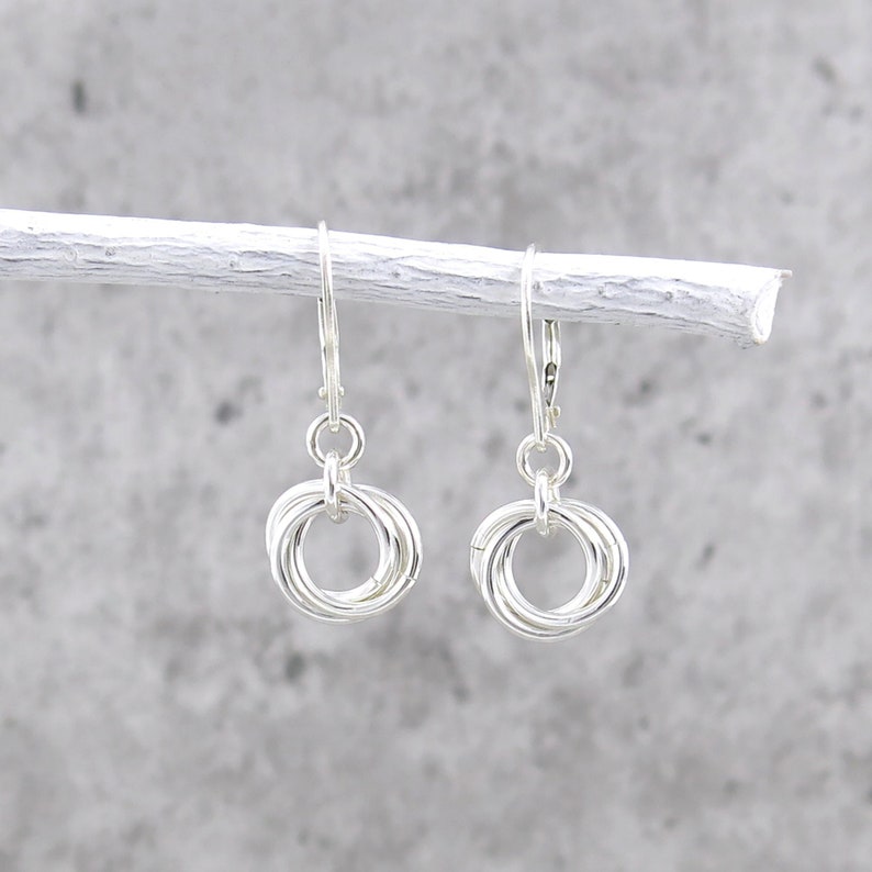May include: A pair of silver-colored earrings. Each earring features a cluster of interlocking rings suspended from a lever-back closure. The earrings are displayed against a light gray background, creating a minimalist aesthetic.