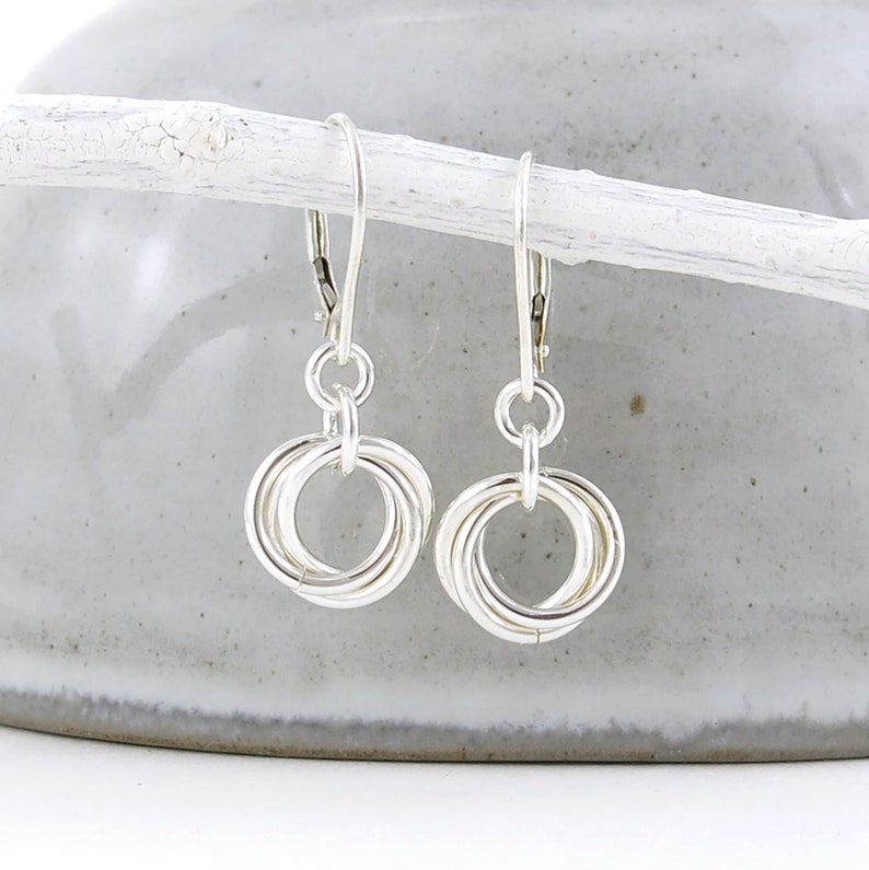 May include: A pair of silver-colored earrings. Each earring features three interlocking rings suspended from a lever-back clasp. The earrings are displayed against a neutral background, highlighting their metallic shine and intricate design.