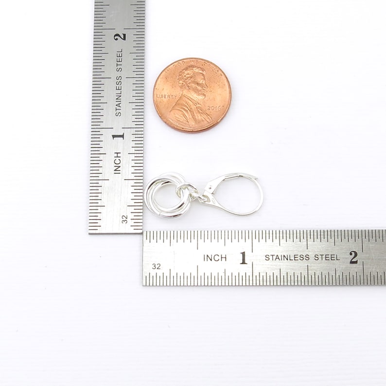 May include: A silver-colored earring with a lever back clasp. The earring features a design of four interlocking circles. A penny is next to the earring for size comparison. The earring is approximately 0.75 inches in length.