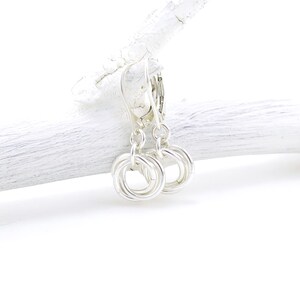 May include: A pair of silver-colored earrings with a circular design. Each earring features three interlocking rings, creating a textured, knotted appearance. The earrings hang from a lever-back clasp. The background is a white, textured surface.