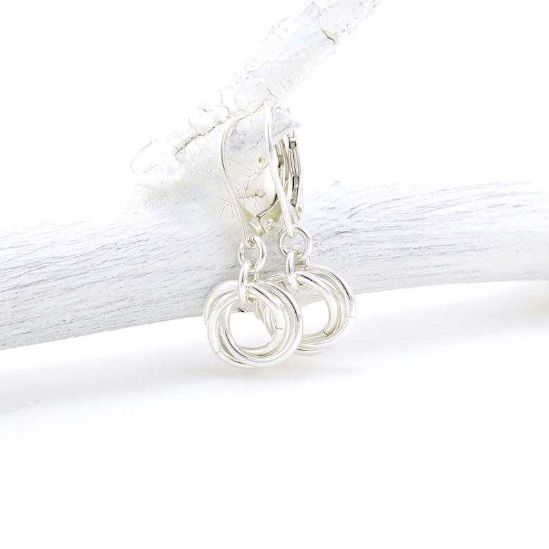 May include: A pair of silver-colored earrings with a circular design. Each earring features three interlocking rings, creating a textured, knotted appearance. The earrings hang from a lever-back clasp. The background is a white, textured surface.