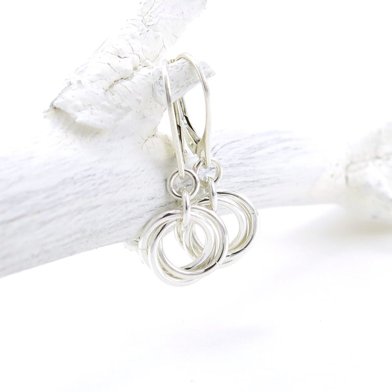 May include: A pair of silver-toned earrings featuring a cluster of interlocking rings. The earrings hang from a lever-back clasp. The jewelry is displayed against a white, textured background, creating a clean and elegant presentation.