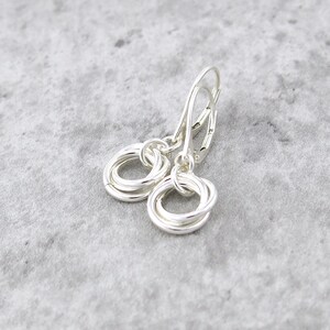 May include: A pair of silver earrings with a lever back closure. Each earring features a cluster of intertwined silver rings, creating a textured, circular design. The earrings are displayed on a light gray surface.