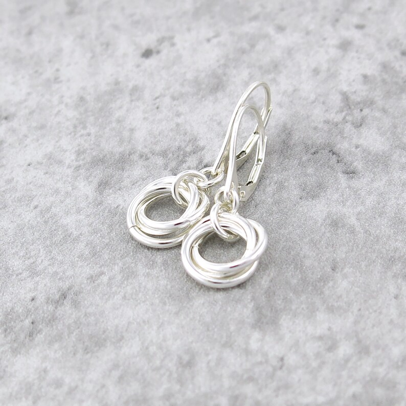 May include: A pair of silver earrings with a lever back closure. Each earring features a cluster of intertwined silver rings, creating a textured, circular design. The earrings are displayed on a light gray surface.