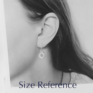 May include: A close-up of a silver earring with a small, dangling design of three interlocking circles. The earring is shown next to an ear for size reference. The text "Size Reference" is at the bottom of the image.