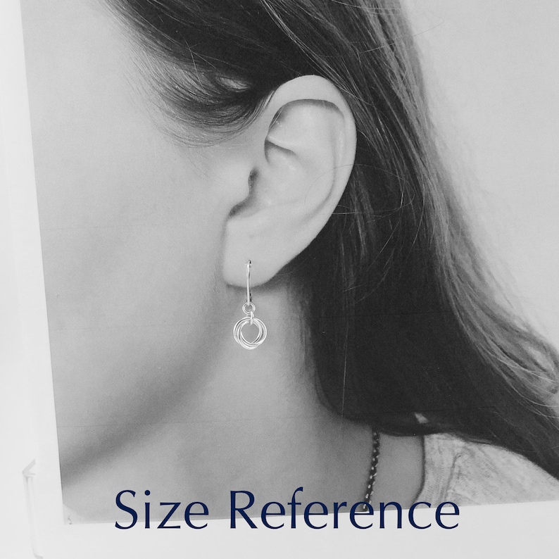 May include: A close-up of a silver earring with a small, dangling design of three interlocking circles. The earring is shown next to an ear for size reference. The text "Size Reference" is at the bottom of the image.