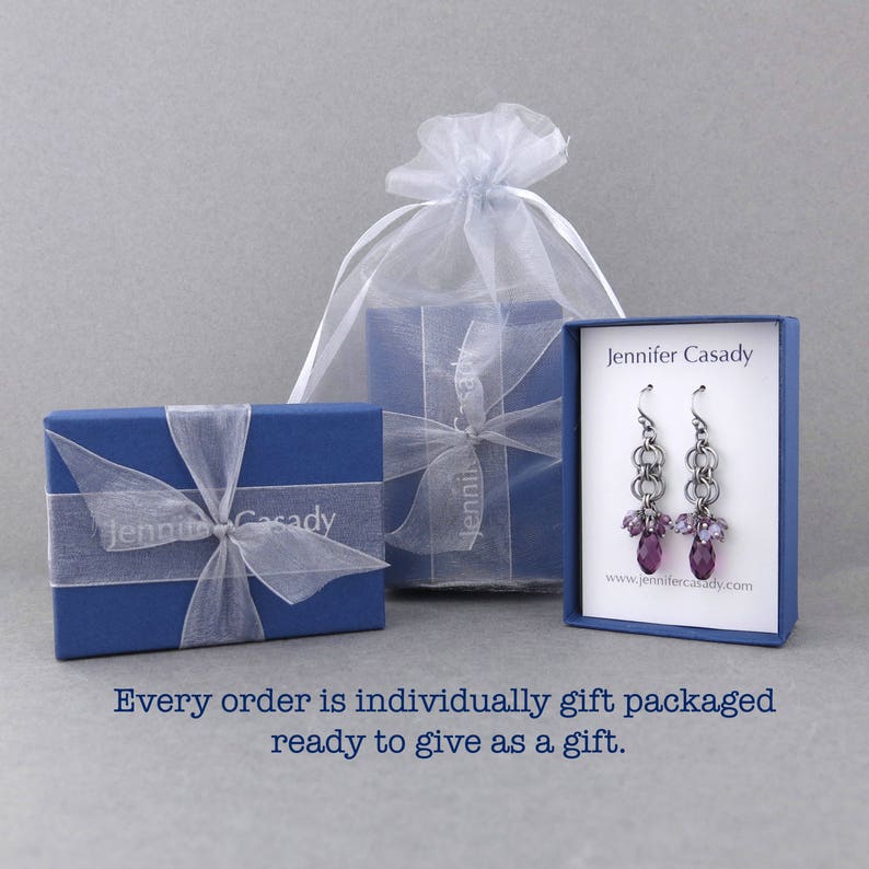 Puede incluir: Un par de pendientes de cristal morado con eslabones de cadena plateados, empaquetados en una caja de regalo azul con una cinta blanca y una bolsa de organza blanca. La caja tiene el texto "Jennifer Casady" impreso en ella.