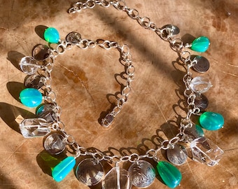 May include: A silver chain necklace with multiple charms, including clear quartz crystals, green gemstones, and silver coins.