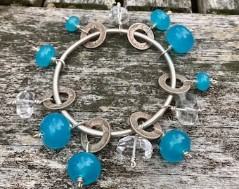 May include: A handmade bracelet featuring a silver metal design with turquoise blue beads, clear crystal accents, and vintage coin details. The bracelet is displayed on a weathered wooden surface.