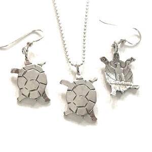 May include: A silver necklace and earrings set featuring a turtle design. The turtle pendant and earrings are detailed with a textured shell pattern.