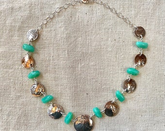 May include: A silver-toned necklace featuring alternating elements of textured, coin-like pendants and light green, oval-shaped beads. The necklace has a delicate chain and a secure clasp. The pendants have a two-tone appearance, with silver and bronze hues.
