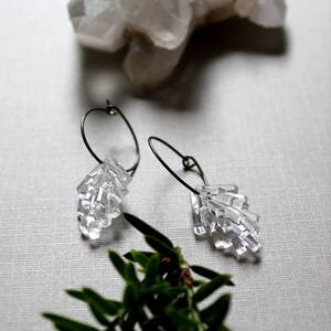May include: A pair of silver hoop earrings with clear crystal geometric dangle charms. The earrings are on a white surface with a small green plant.