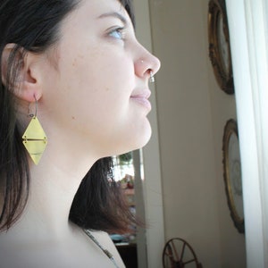 May include: A woman wearing a gold geometric earring. The earring is a double-sided triangle with a line across the center.