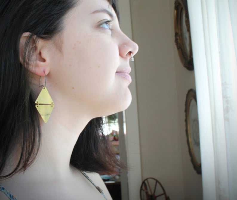 May include: A woman wearing a gold geometric earring. The earring is a double-sided triangle with a line across the center.