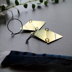 May include: A pair of gold geometric earrings with black wire accents. The earrings are on a gray surface with a feather.