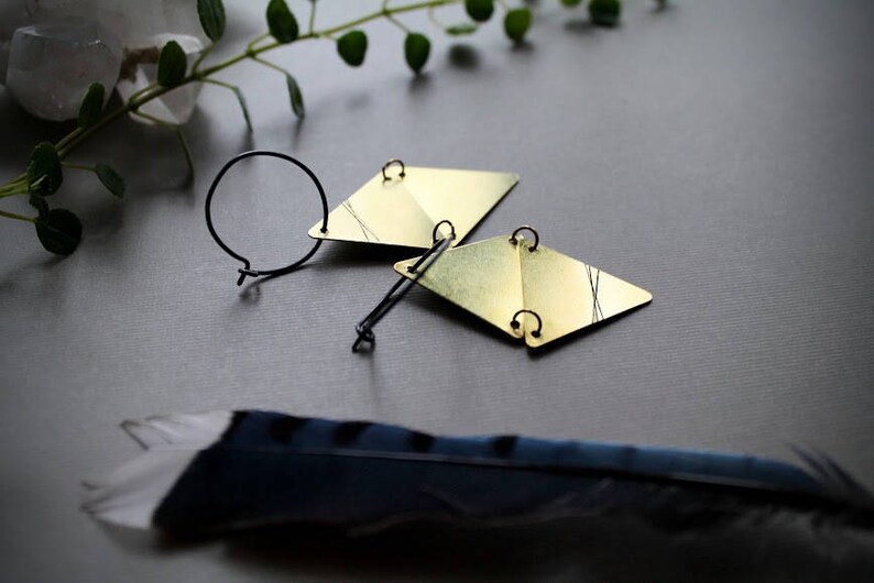 May include: A pair of gold geometric earrings with black wire accents. The earrings are on a gray surface with a feather.