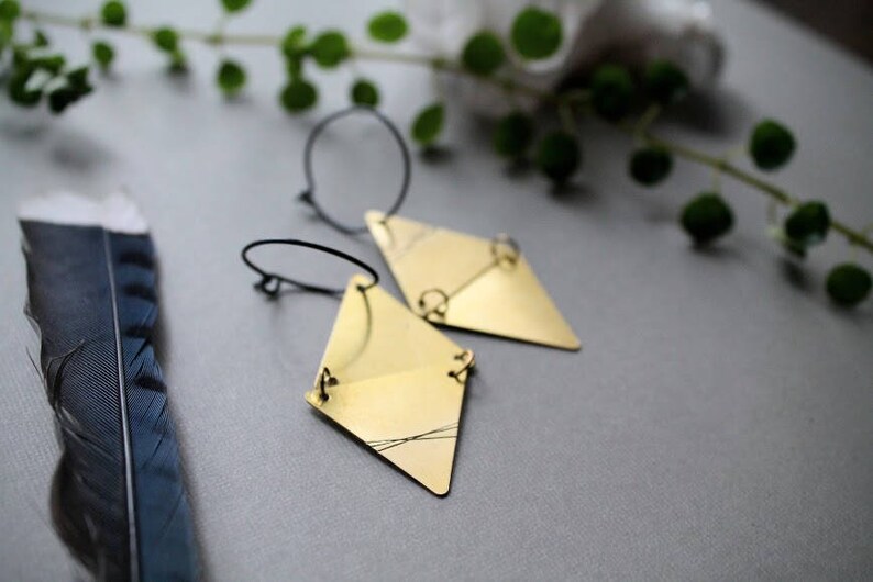 May include: A pair of gold-toned geometric earrings with a diamond shape. The earrings are suspended from thin black hoops.