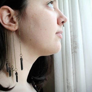 May include: A woman with long black hair wearing a long, dangling ear cuff with multiple black crystal charms. The ear cuff is attached to her ear with a small hoop.