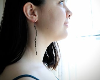 May include: A person wearing long, dark hair and a teal top with a long, thin, black earring. The earring is made of a series of small, connected circles.