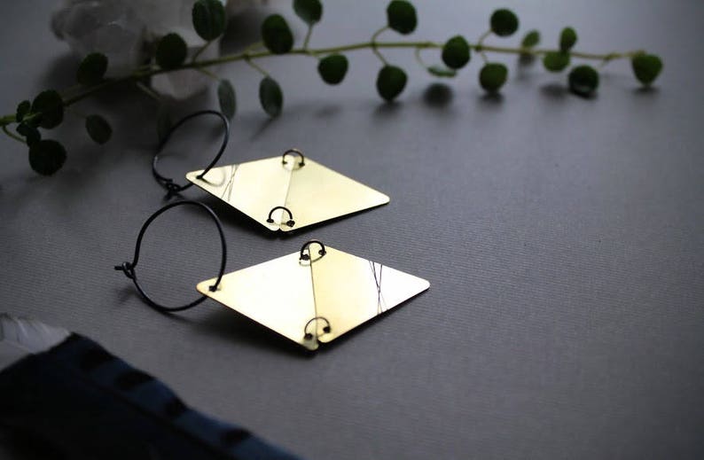 May include: A pair of gold-toned earrings with black hoops and diamond-shaped pendants. The earrings are on a gray surface.