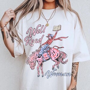 Puede incluir: Camiseta color crema con un diseño de una vaquera montando un caballo rosa y sosteniendo un libro. El texto "Well Read Woman" está escrito en rosa y azul en la camiseta.