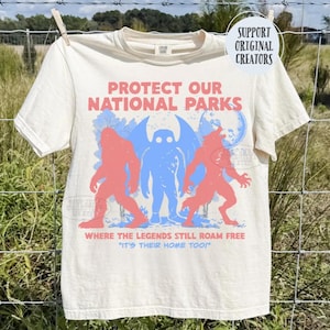 Può includere: Una t-shirt a maniche corte color crema con il testo rosso PROTECT OUR NATIONAL PARKS e WHERE THE LEGENDS STILL ROAM FREE. La grafica mostra sagome rosse e blu di creature leggendarie con il testo blu IT'S THEIR HOME TOO!