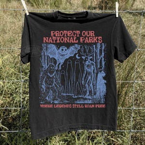 May include: A black t-shirt featuring a blue graphic print of a forest scene with a bat, a sasquatch, ghosts, and a deer-like creature. The text reads "PROTECT OUR NATIONAL PARKS" at the top and "WHERE LEGENDS STILL ROAM FREE" at the bottom in red.