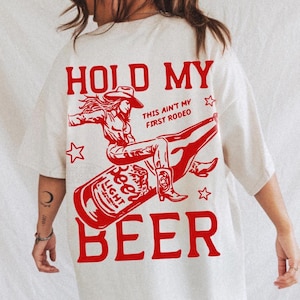 Op de afbeelding: Een gebroken wit t-shirt met een grote rode afbeelding op de achterkant. Het ontwerp toont een persoon met een cowboyhoed die op een bierfles rijdt, met de tekst "HOLD MY BEER" en "THIS AIN'T MY FIRST RODEO" en kleine sterren.