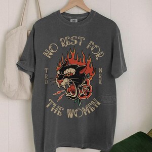 May include: Charcoal grey t-shirt with a retro graphic. The design includes a black panther with red flames, a rose, and the text "NO REST FOR THE WOMEN". The t-shirt is displayed on a wooden hanger.