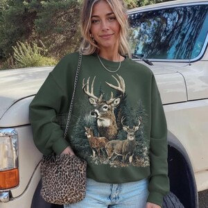 May include: An olive green sweatshirt featuring a detailed graphic of a deer family in a forest setting. The sweatshirt is paired with light blue jeans and a leopard print purse. The background includes a vintage white truck and trees.