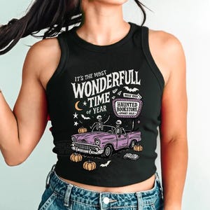 May include: Black tank top with a Halloween-themed graphic. The design features the text "It's the Most Wonderfull Time of Year" with a vintage car carrying skeletons, pumpkins, and the words "Haunted Bookstore".