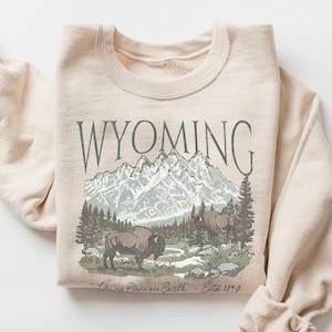 Könnte beinhalten: Beiges Sweatshirt mit einem Wyoming-Motiv. Das Design zeigt Berge, Bäume und Bisons, mit dem Wort "WYOMING" darüber. Der Text "Like Heaven on Earth - Estd 1890" ist unter der Grafik aufgedruckt. Das Sweatshirt ist gefaltet.