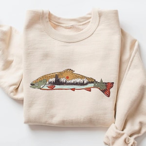 May include: A cream-coloured sweatshirt with a detailed illustration of a fish containing a mountain and sunset scene. The fish design includes shades of brown, green, red, and orange. The sweatshirt has a crew neck and long sleeves.