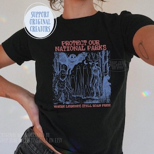 Può includere: Una t-shirt nera a maniche corte con una stampa grafica in blu e rosso. Il testo recita PROTECT OUR NATIONAL PARKS e WHERE LEGENDS STILL ROAM FREE. L'illustrazione raffigura un pipistrello, un sasquatch, dei fantasmi e una creatura simile a un cervo.