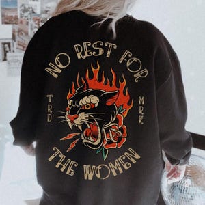 May include: Black sweatshirt with a graphic of a panther's head engulfed in flames, a red rose, and the text "NO REST FOR THE WOMEN." The sweatshirt is paired with an orange cap.